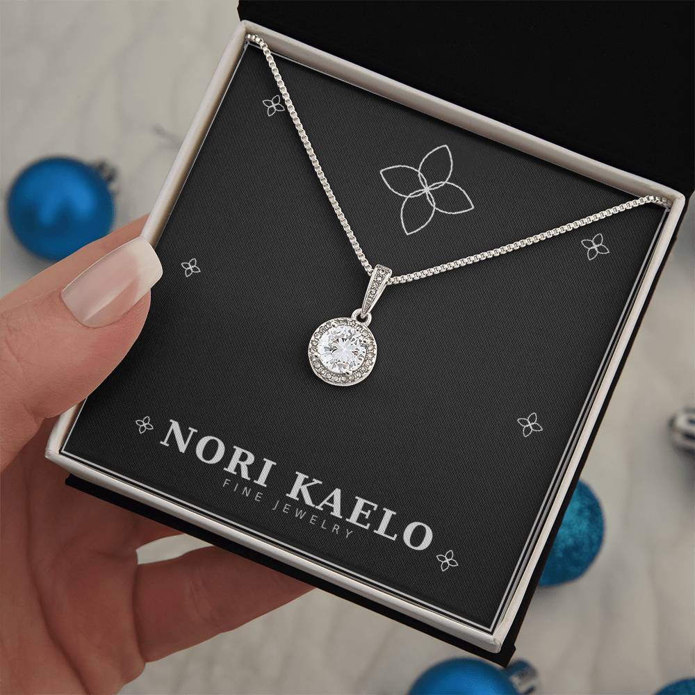 A hand holding an open Nori Kaelo Fine Jewelry box displaying a halo pendant necklace, with blue holiday ornaments in the background.