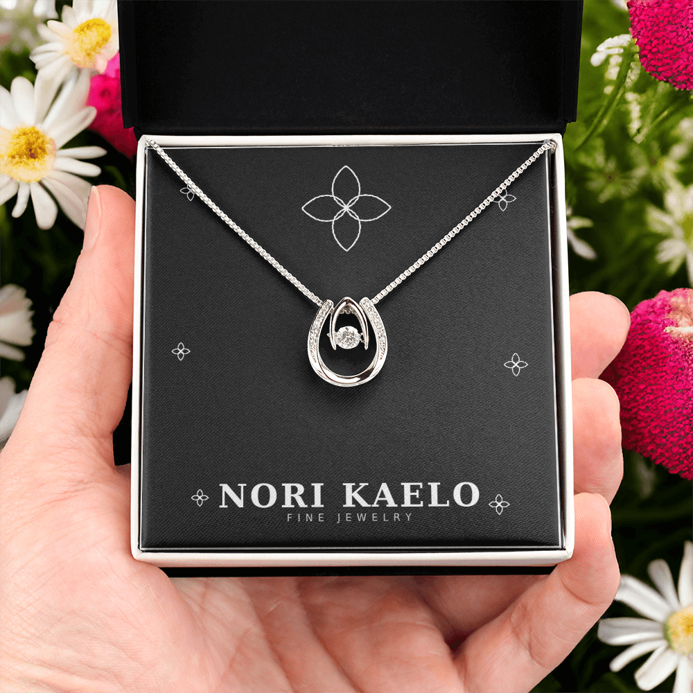 A hand holding an open Nori Kaelo Fine Jewelry box displaying a dancing stone pendant necklace in white gold, styled with flowers in the background.