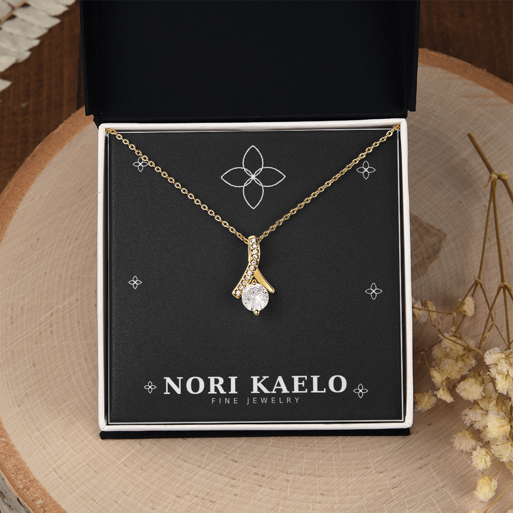 Ribbon pendant necklace in yellow gold, displayed on a gift box with a rustic, botanical styling in the background.