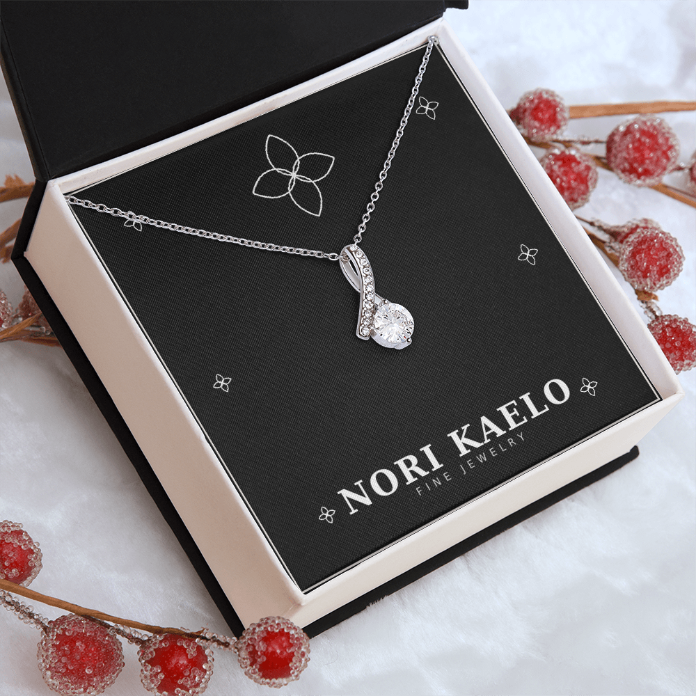 Ribbon pendant necklace in white gold, displayed on a gift box styled with flowers in the background.