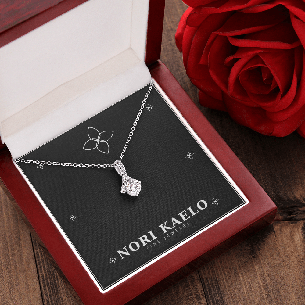 Ribbon pendant necklace in white gold, displayed in an open mahogany gift box, styled with a red rose in the background.