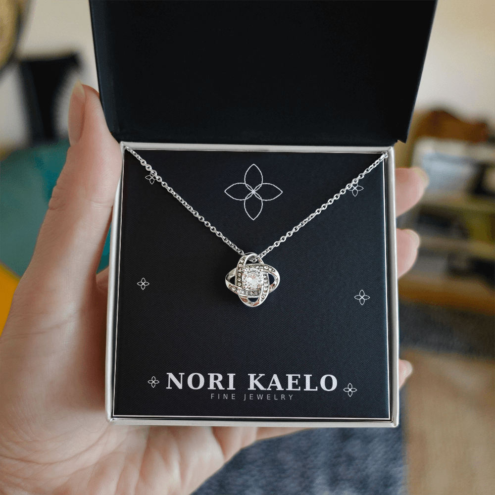 A hand holding an open Nori Kaelo Fine Jewelry box displaying a love knot pendant necklace in white gold.