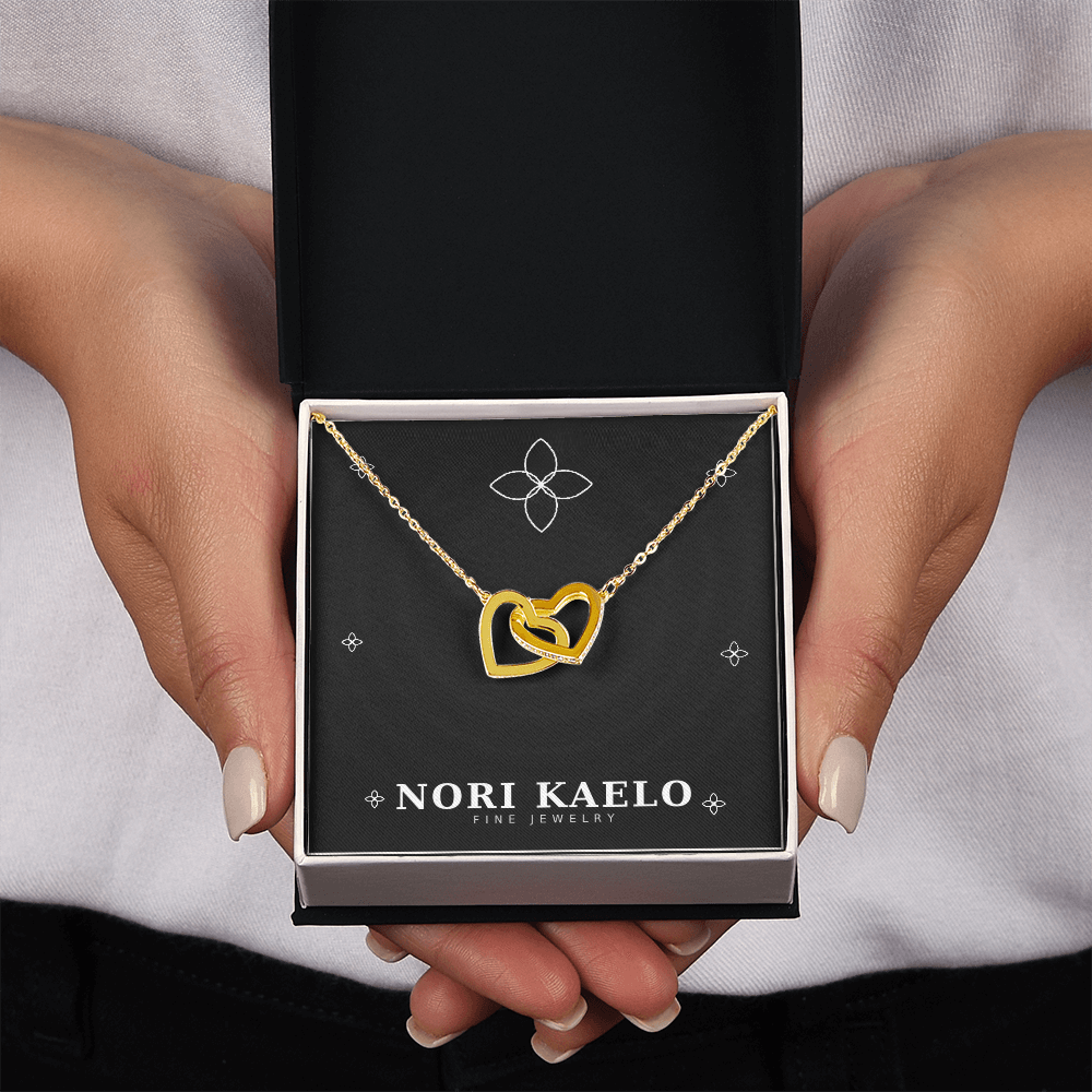 Two hands holding an open Nori Kaelo Fine Jewelry box displaying an interlocking hearts pendant necklace in yellow gold.