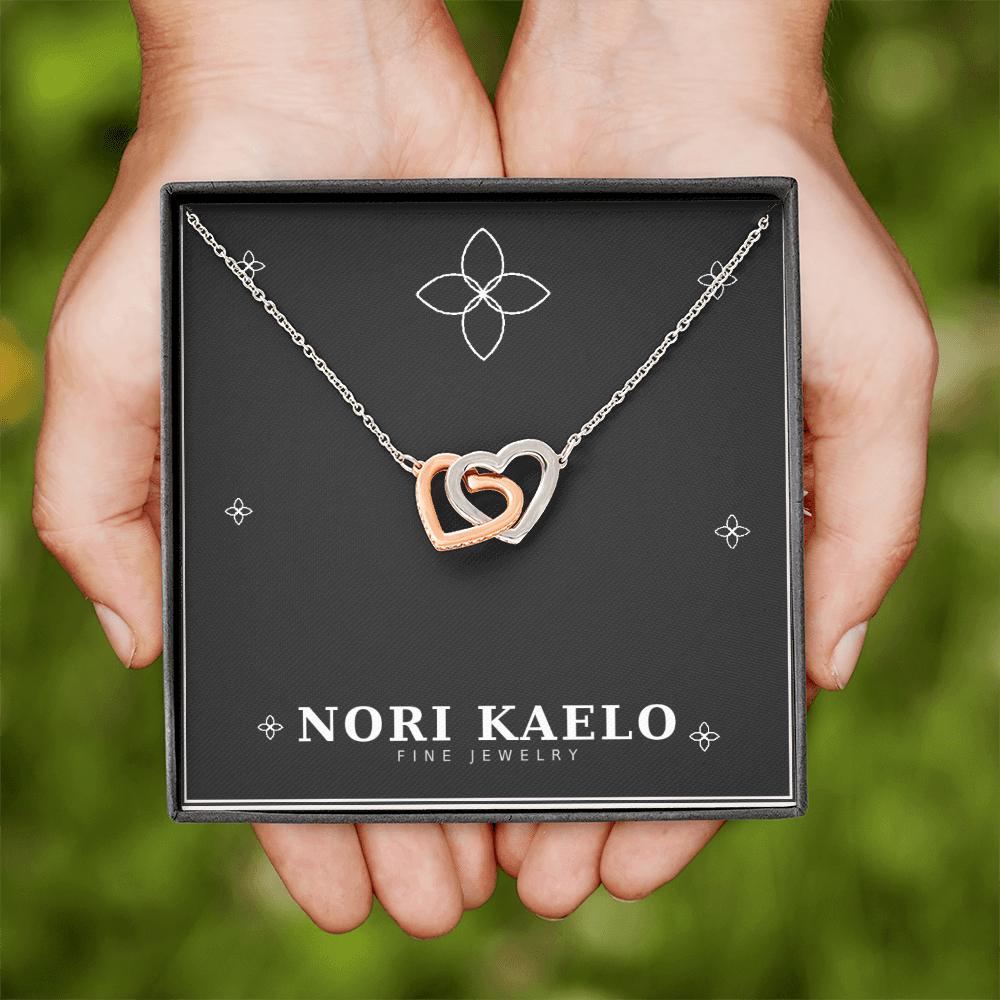 Two hands holding an open Nori Kaelo Fine Jewelry box displaying an interlocking hearts pendant necklace in rose gold and white gold, outdoors with greenery in the background.