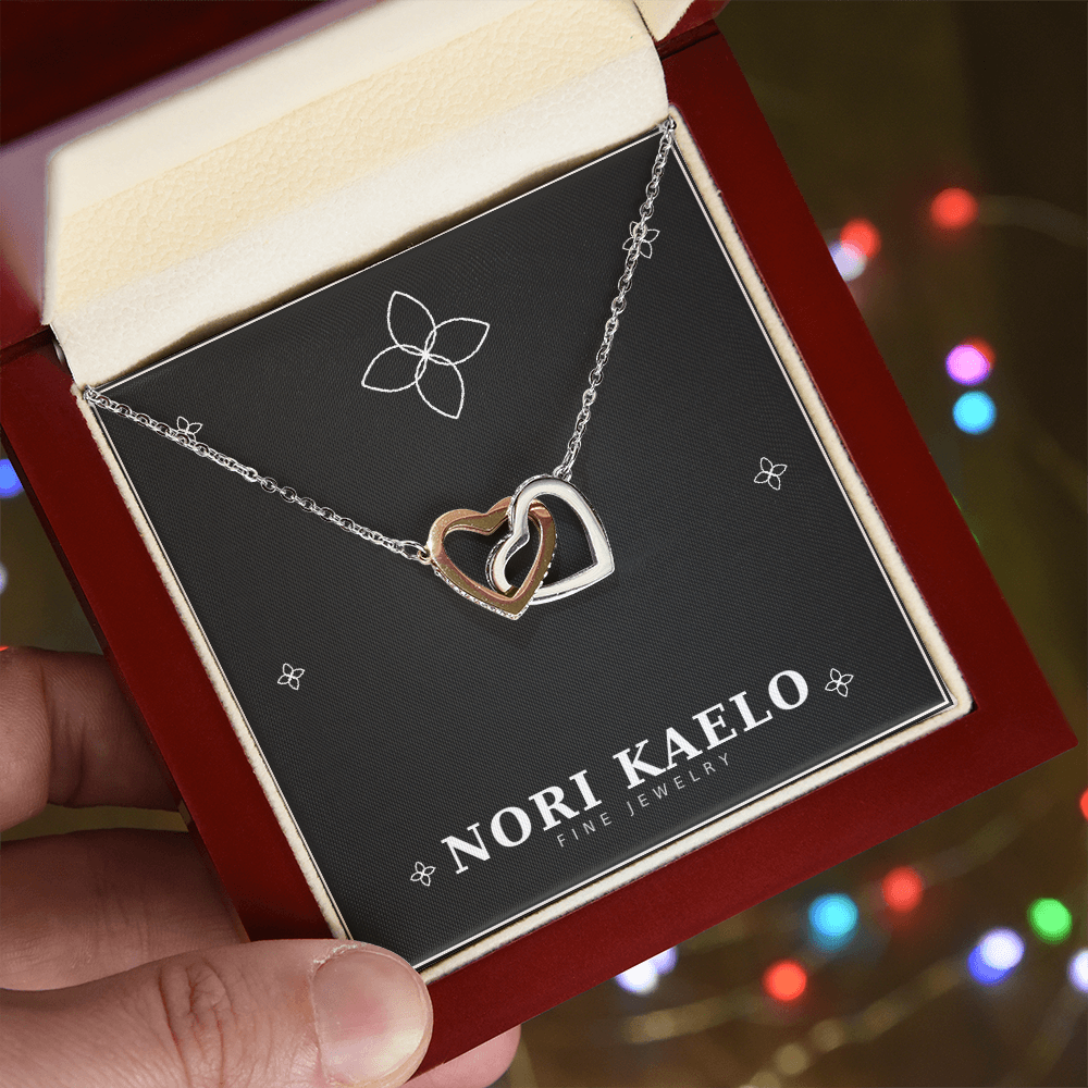 Interlocking hearts pendant necklace in rose gold and white gold, displayed in a mahogany gift box, with string lights in the background.