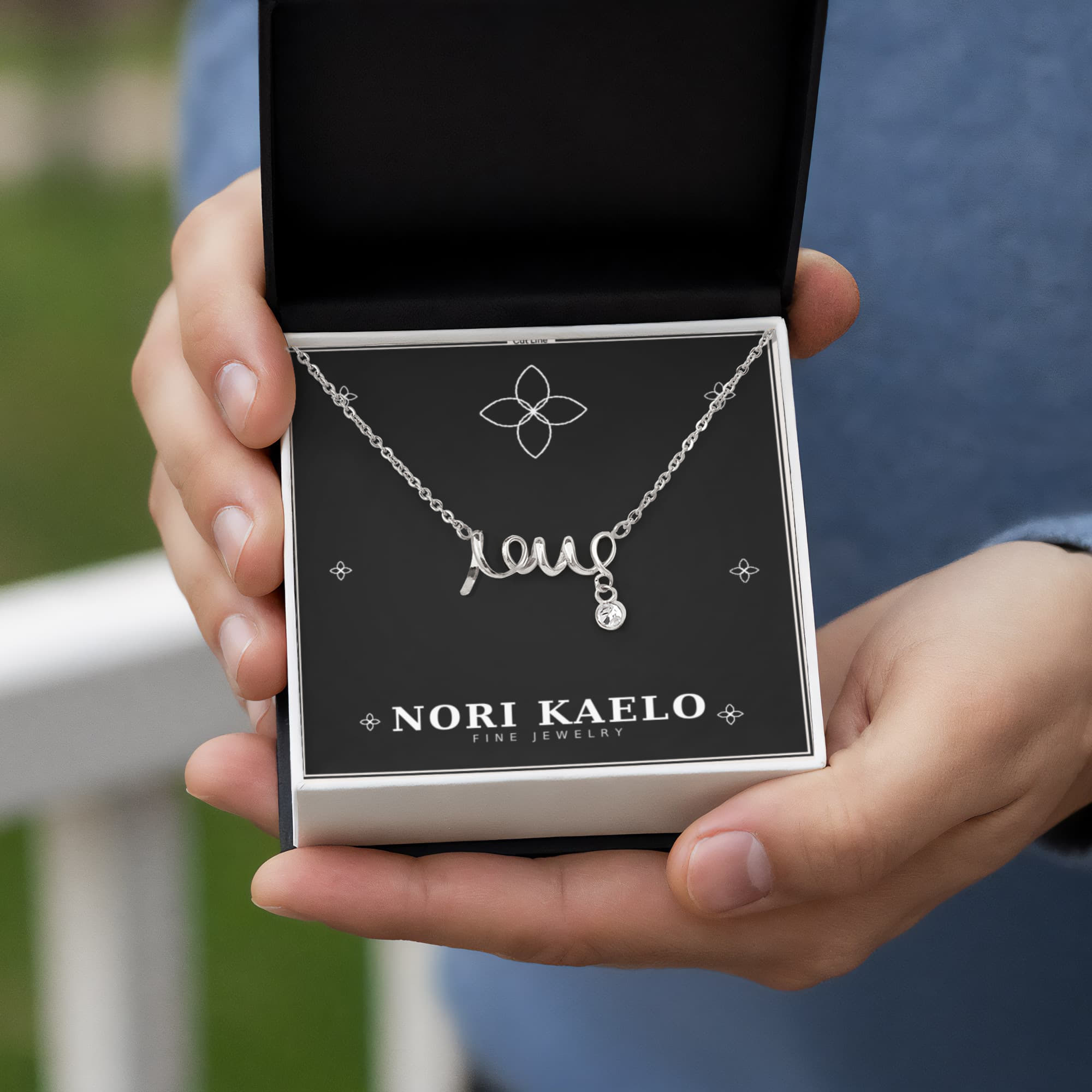 Scripted Love Necklace by Nori Kaelo, silver script pendant held in an open gift box outdoors